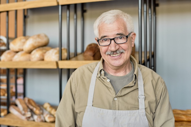 Retrato de panadero de edad feliz en anteojos y delantal de pie contra el puesto con pan en la tienda