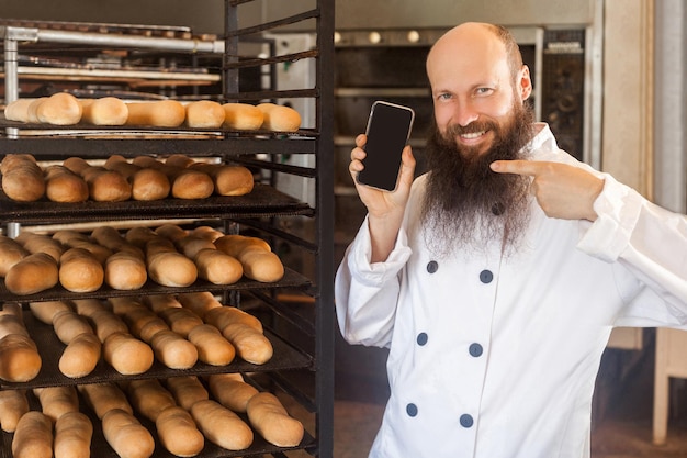 Retrato de panadero de negocios adulto satisfecho con barba en uniforme blanco de pie en la panadería y esperando un pedido en línea por teléfono, señalando con el dedo al teléfono con una sonrisa dentuda Interior, mirando a la cámara