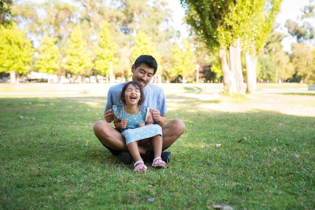 Retrato de papá feliz divirtiéndose con su pequeña hija en el parque. Joven asiático sentado en la hierba verde jugando con una chica que se ríe haciéndole cosquillas en las manos. Concepto de actividad de ocio de paternidad y verano.