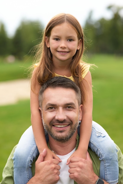 Retrato de papá feliz sosteniendo a su linda hijita sobre los hombros y sonriendo a la cámara mientras