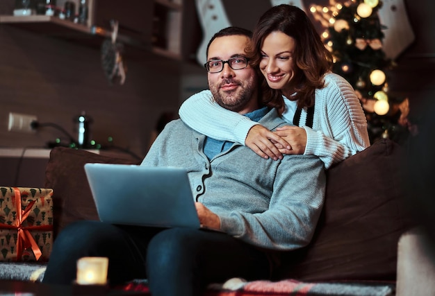 Retrato de una pareja adorable con una laptop, una mujer encantadora con sombrero de Santa abrazando a su hombre y mirando la cámara. Mujer celebrando la Nochebuena.