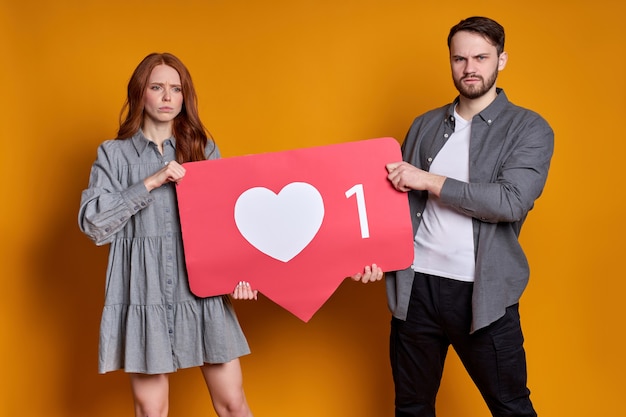 Retrato de pareja alegre en ropa de fiesta con corazón como icono, recomendando hacer clic en el botón de redes sociales