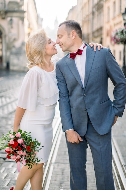 Retrato de pareja amorosa de cerca, hombre guapo de 50 años en traje y pajarita, de pie con su mujer bonita en vestido blanco con ramo de flores, en el fondo de la ciudad vieja