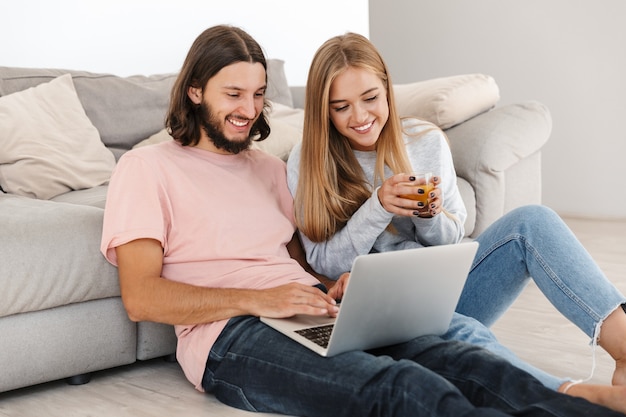 Retrato de pareja amorosa joven feliz optimista cerca de sofá usando computadora portátil en el interior de casa.