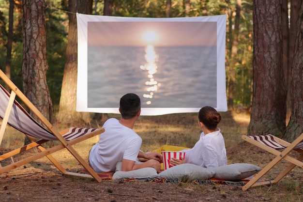 Retrato de una pareja amorosa relajada tirada en el bosque con un proyector de techo viendo una película romántica disfrutando de la puesta de sol en la pantalla y la hermosa naturaleza en el bosque