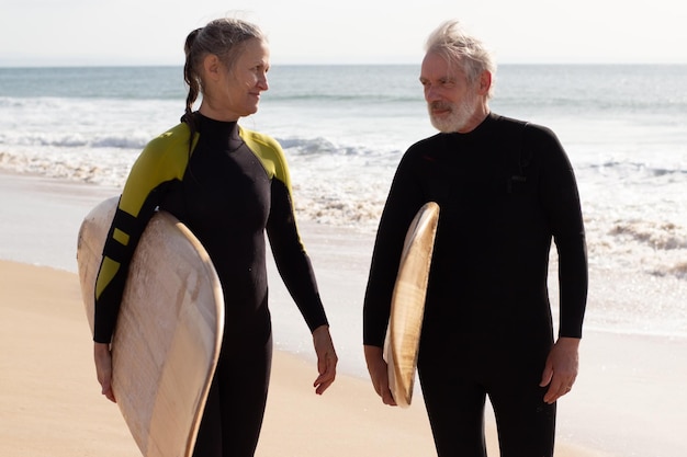 Retrato de pareja anciana lista para montar en tablas. Hombre y mujer deportivos en trajes de neopreno caminando por la orilla del mar con tablas de surf, preparándose para surfear y hablar. Deporte, actividad de ocio del concepto de personas mayores.