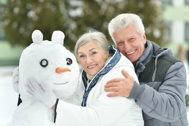 Retrato de pareja de ancianos haciendo muñeco de nieve y muestra los pulgares para arriba en invierno