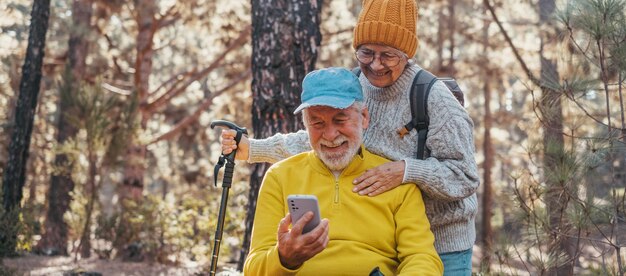 Retrato de una pareja de ancianos lindos usando el teléfono juntos en el bosque con árboles a su alrededor usando el teléfono móvil al aire libre divirtiéndose y disfrutando