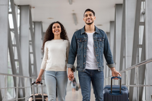 Retrato de una pareja árabe milenaria positiva caminando en el aeropuerto con equipaje