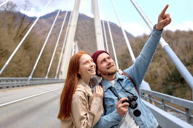 Retrato de pareja caucásica en ropa casual caminando sobre el puente, en contemplación de montañas, paisajes alrededor. Dedo acusador masculino guapo al lado, mostrando algo a la novia