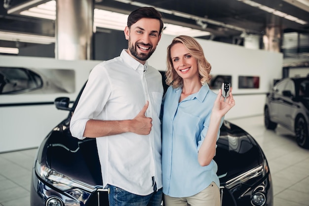 Retrato de una pareja feliz sosteniendo llaves con un auto negro nuevo en el fondo del salón del concesionario