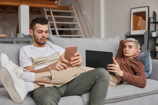 Retrato de una pareja gay contemporánea usando computadoras mientras se relajan en el sofá juntos en el interior de una casa mínima, espacio de copia