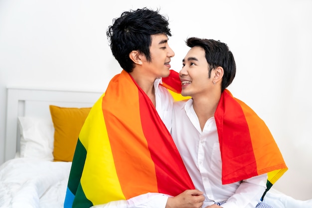 Foto retrato de pareja de homosexuales asiáticos abrazo y mano con bandera de orgullo en el dormitorio. concepto lgbt gay.