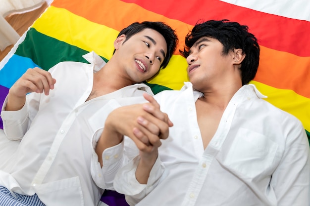 Retrato de pareja de homosexuales asiáticos abrazo y mano con bandera de orgullo en el dormitorio. Concepto LGBT gay.