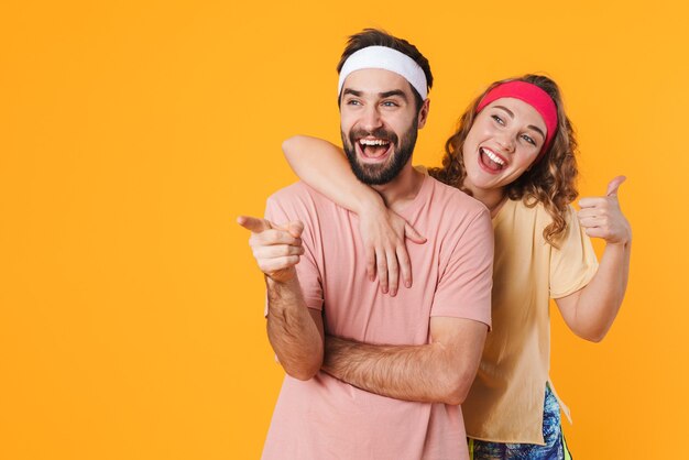 Retrato de pareja joven atlética con diademas sonriendo y gesticulando con el pulgar hacia arriba aislado sobre la pared amarilla