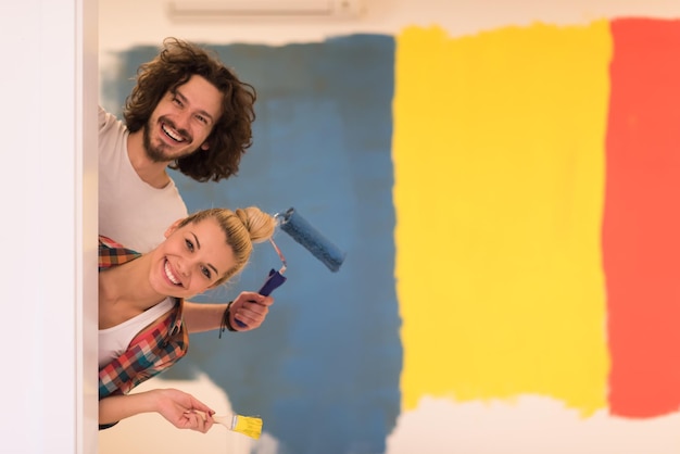 retrato de una pareja joven feliz y sonriente pintando la pared interior de una casa nueva asomándose desde detrás de la pared