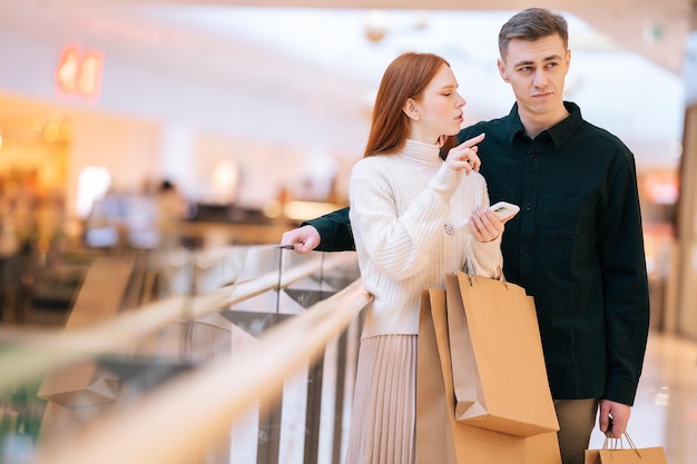 Retrato de una pareja joven preocupada que busca una tienda en el centro comercial aplicación de navegación en el teléfono móvil Hombre y mujer estresados que usan un teléfono inteligente de pie con bolsas de papel con compras en la tienda