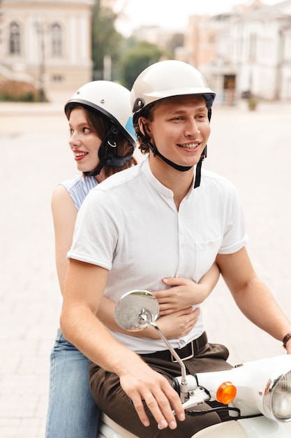 Retrato de una pareja joven sonriente en cascos montando en una moto juntos en la calle de la ciudad