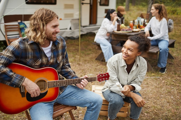 Retrato de pareja joven tocando la guitarra mientras acampa con amigos en el espacio de copia del bosque