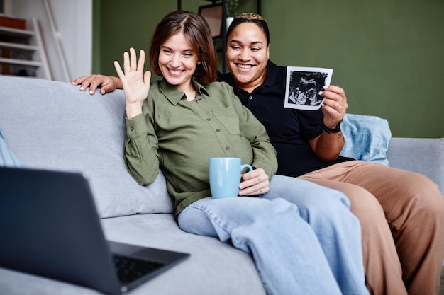 Retrato de una pareja lesbiana sonriente esperando un bebé y mostrando una imagen de ultrasonido a la familia a través de un video c