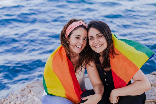 Foto retrato de una pareja de lesbianas sonrientes con una bandera arco iris sentadas junto al mar