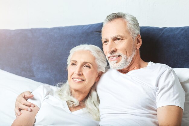 Retrato de una pareja mayor sonriente viendo la televisión en casa sentada en la cama