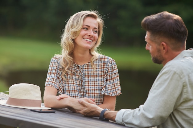 Retrato de pareja romántica tomados de la mano y mirarse con amor mientras está sentado en la mesa al aire libre junto al lago