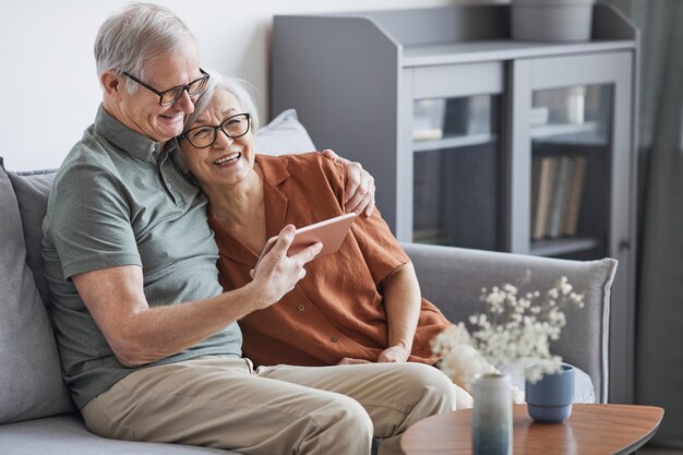 Retrato de pareja senior moderna usando tableta en casa y sonriendo felizmente mientras está sentado en el sofá en un interior mínimo, espacio de copia