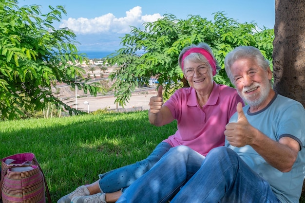 Retrato de pareja senior sonriente sentada en el césped en un parque público mirando a la cámara con los pulgares hacia arriba Pareja romántica disfrutando del tiempo libre y la jubilación