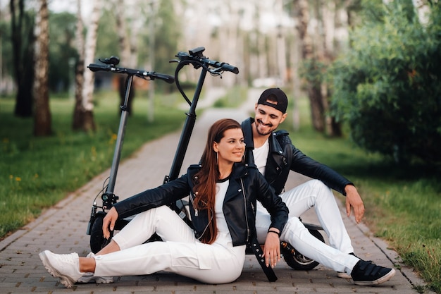 Retrato de una pareja sentada cerca de scooters eléctricos, disfrutando juntos de tiempo en la naturaleza, dos amantes en scooters eléctricos