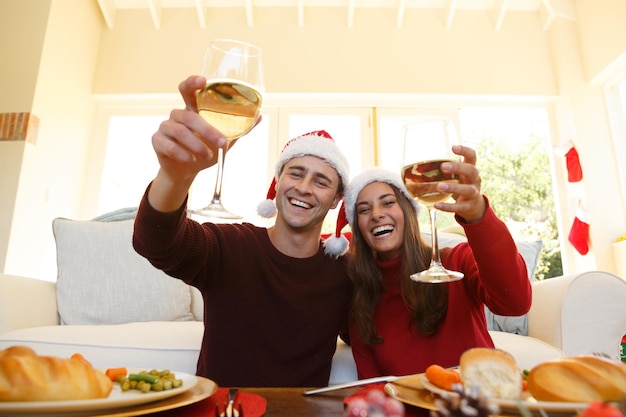 Retrato de una pareja con sombrero de Papá Noel brindando mirando a la cámara mientras está sentado en la sala de estar de casa. concepto de celebración de tradición navideña