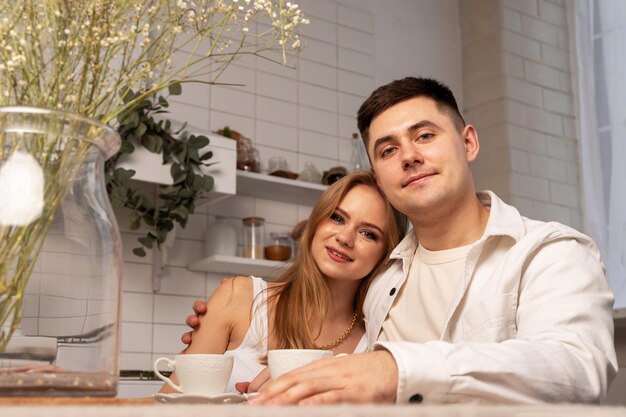 Retrato de una pareja sonriente sentada en la mesa de la cocina con tazas de té o café y flores secas han