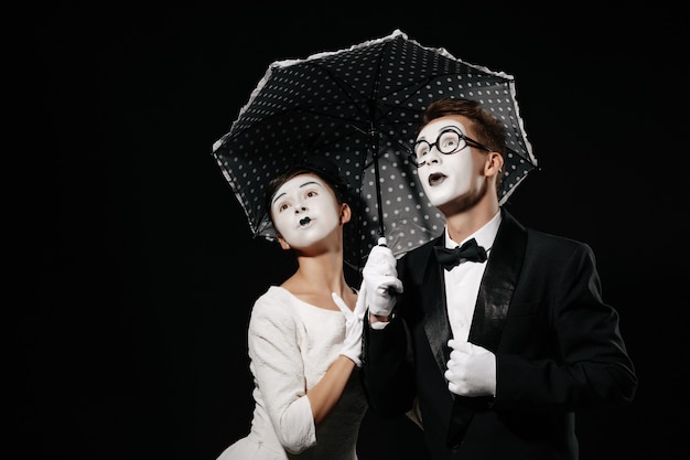 Retrato de pareja sorprendida mimo con paraguas sobre fondo negro. hombre en esmoquin y gafas y mujer en vestido blanco