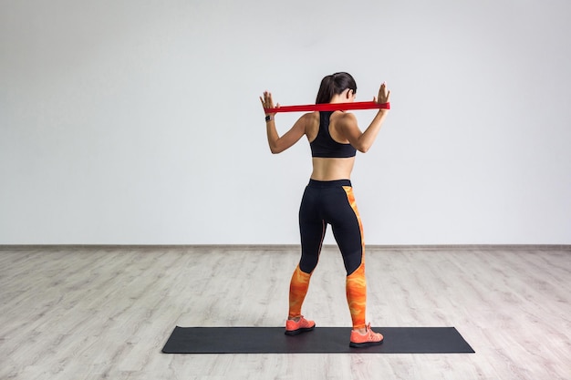 El retrato de la parte trasera de una mujer deportiva vestida con top negro y polainas naranjas realiza ejercicios para los músculos de la espalda y las manos, entrena con una banda de resistencia sobre fondo blanco. Interior