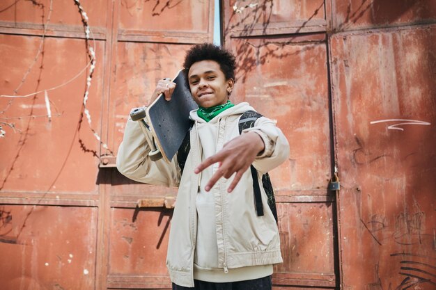 Retrato de un patinador afroamericano sosteniendo patineta y gesticulando a la cámara en el área urbana