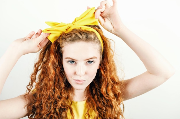 Retrato de una pelirroja rizada con una camiseta amarilla endereza el lazo amarillo en la cabeza mientras está de pie en el fondo blanco