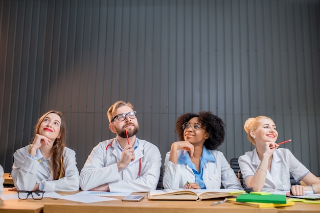 Retrato de un pensativo grupo multiétnico de estudiantes de medicina en uniforme sentados juntos en una fila en el escritorio en el aula moderna