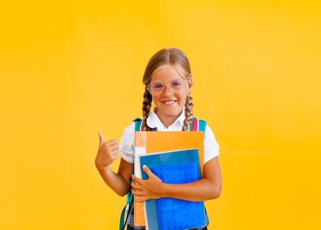 Retrato de una pequeña colegiala sonriente con mochila sosteniendo portátil sobre fondo amarillo