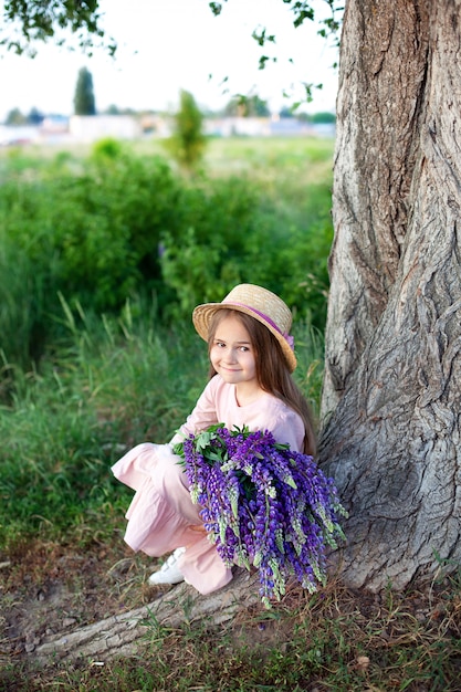 Retrato de pequeña morena sonriente con cabello largo sentado cerca de un árbol con altramuces púrpuras