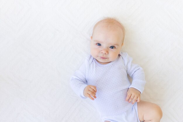 Retrato de una pequeña niña de 6 meses en un traje blanco acostada de espaldas sobre una cama blanca, espacio para texto