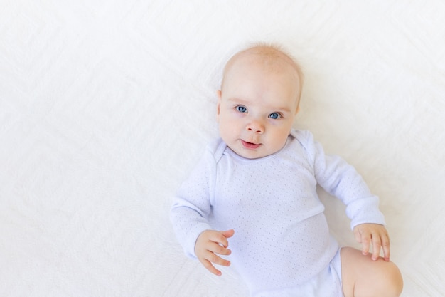 Retrato de una pequeña niña de 6 meses en un traje blanco acostada de espaldas sobre una cama blanca