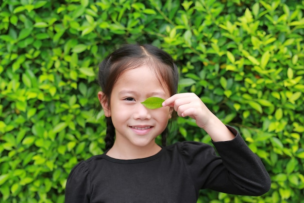 Retrato de la pequeña niña asiática sonriente que sostiene una hoja verde que cierra el ojo derecho