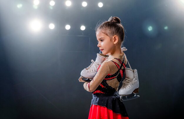 Retrato de pequeña patinadora feliz con patines en sus manos en la arena de hielo, fondo oscuro