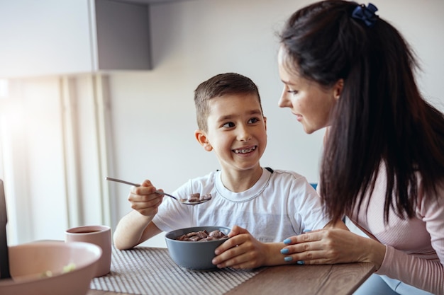 Retrato pequeño adolescente comiendo cereales con leche en kichen y mirando a la madre con una sonrisa