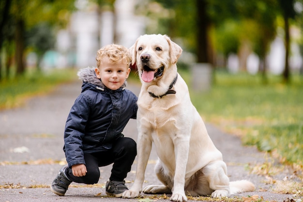 Retrato del pequeño bebé caucásico lindo adorable que se sienta con el perro en parque afuera. Niño sonriente que sostiene el animal doméstico animal doméstico. Concepto de infancia feliz