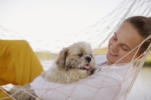 Retrato de perfil de una atractiva mujer caucásica sonriente con camisa blanca yace en una hamaca con un perro pequinés en la orilla del río feliz fin de semana en la naturaleza con su mascota favorita