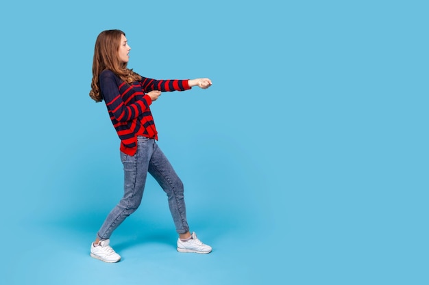 Retrato de perfil completo de una mujer con un suéter de estilo casual a rayas de pie en ataque o tirando de las manos y mirando hacia adelante Foto de estudio interior aislada en fondo azul