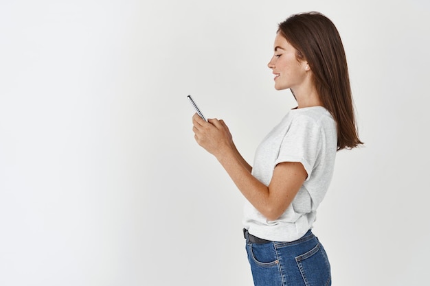 Retrato de perfil de hermosa mujer morena usando teléfono móvil, leyendo mensajes o navegando en las redes sociales, feliz de pie contra la pared blanca.