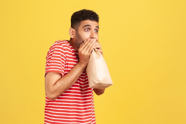 Retrato de perfil hombre asustado con camiseta roja a rayas exhalando e inhalando en un paquete usando una bolsa de papel para mejorar el bienestar superando el miedo al estrés Foto de estudio interior aislada en fondo amarillo