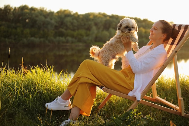 Retrato de perfil de una joven adulta sonriente y feliz con camisa blanca y su perro pequinés sentado al atardecer cerca del río y jugando con su linda mascota durante las vacaciones